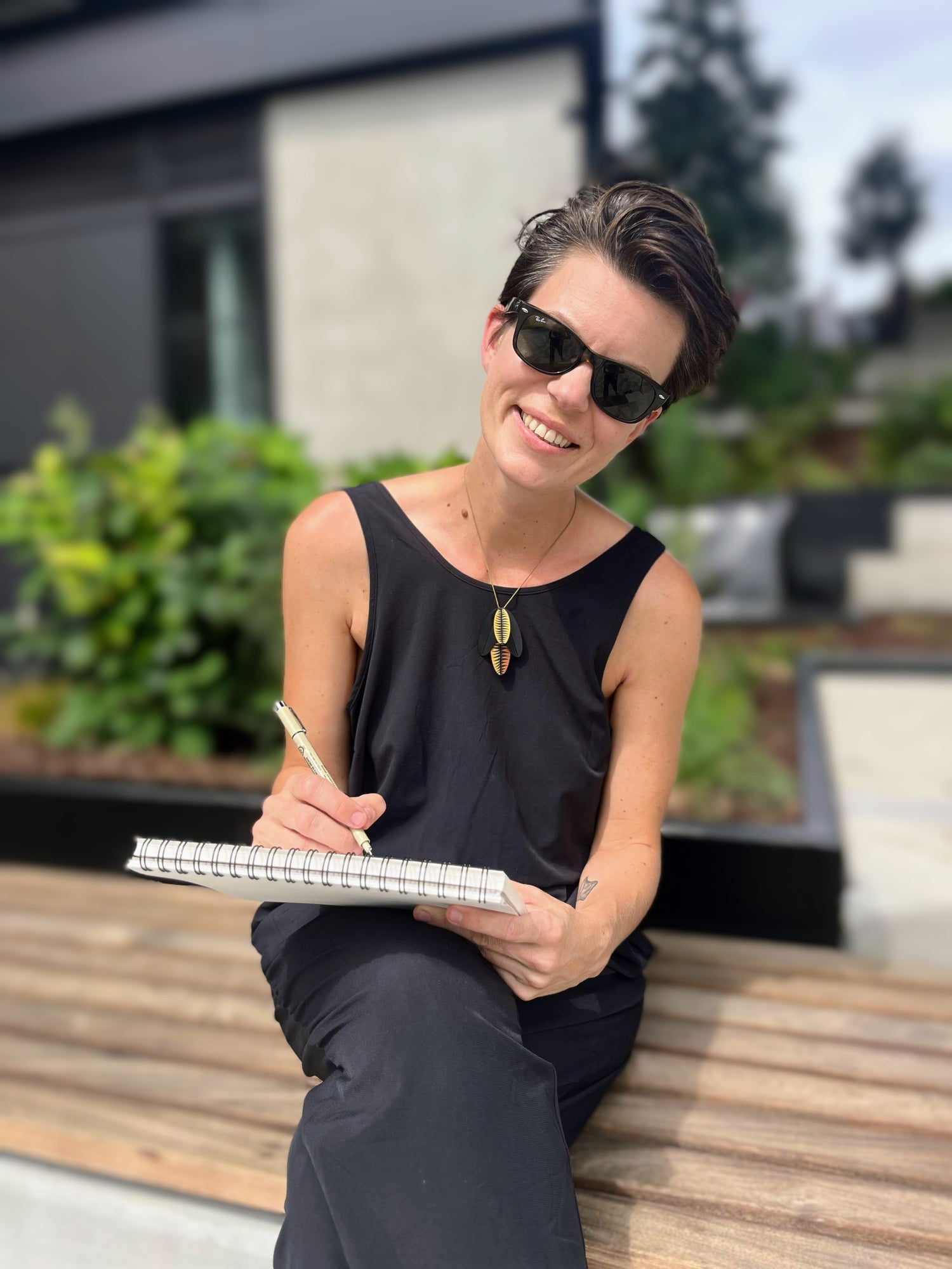 Sara, the artist behind Swil Arts, holding a pencil and notebook. She is wearing dark glasses and is looking at the camera with a big smile. Sara is wearing a black, sleeveless onesie.