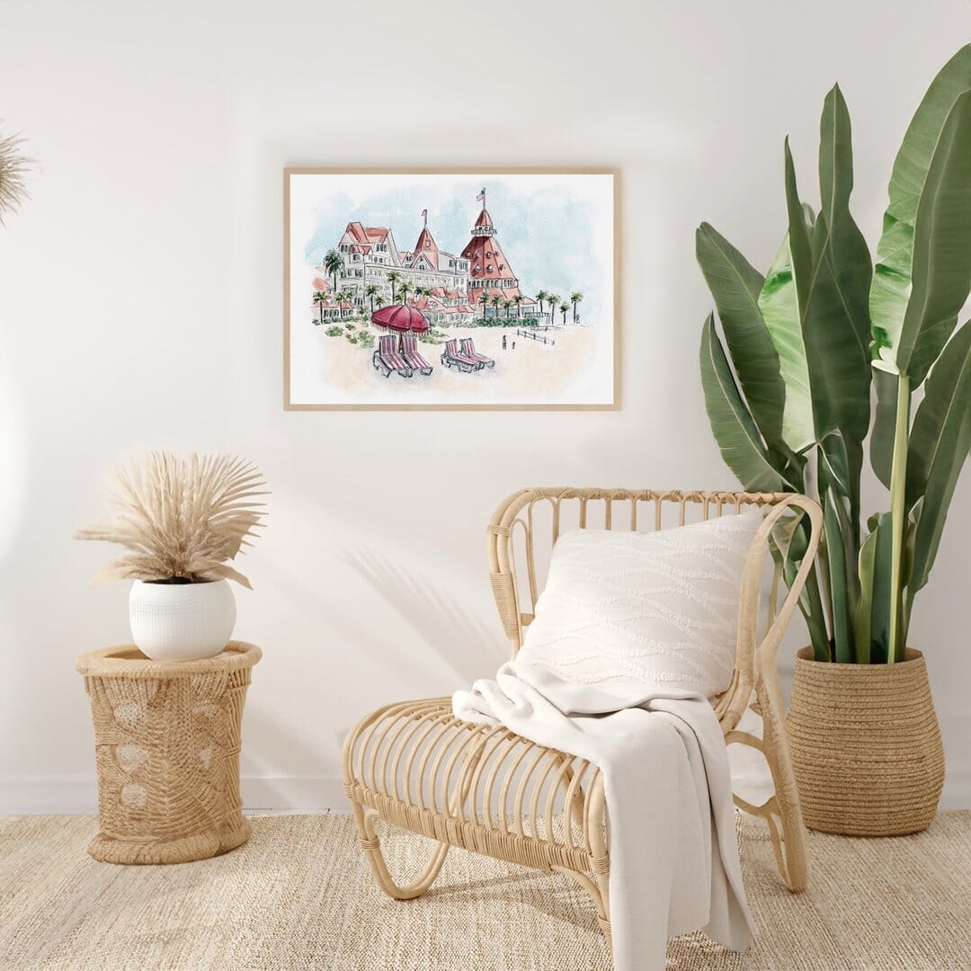 Framed watercolor print of Hotel del Coronado displayed in a bright, airy room with a coastal-inspired aesthetic. The beachy artwork complements the natural rattan chair, soft throw blanket, and neutral decor, creating a relaxing and inviting space. Perfect for beach lovers and San Diego enthusiasts