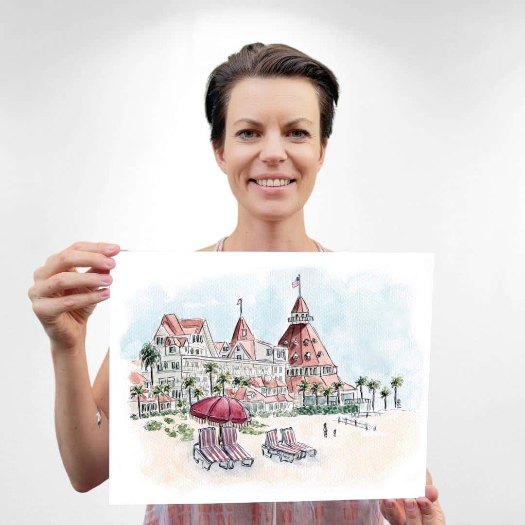 Artist holding a watercolor print of the iconic Hotel del Coronado on the beach, with striped lounge chairs and umbrellas in the foreground. Perfect coastal wall art for fans of historic landmarks and beach-inspired decor. Unique San Diego art piece ideal for travel enthusiasts and collectors