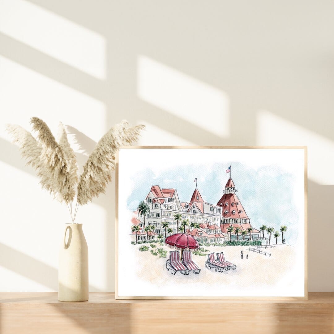 Framed watercolor print of the historic Hotel del Coronado in San Diego, displayed on a wooden shelf with a vase of pampas grass. Coastal California art by Swil Arts, featuring the iconic red turret and beach scene. Perfect beach house wall decor with vintage architecture and natural styling
