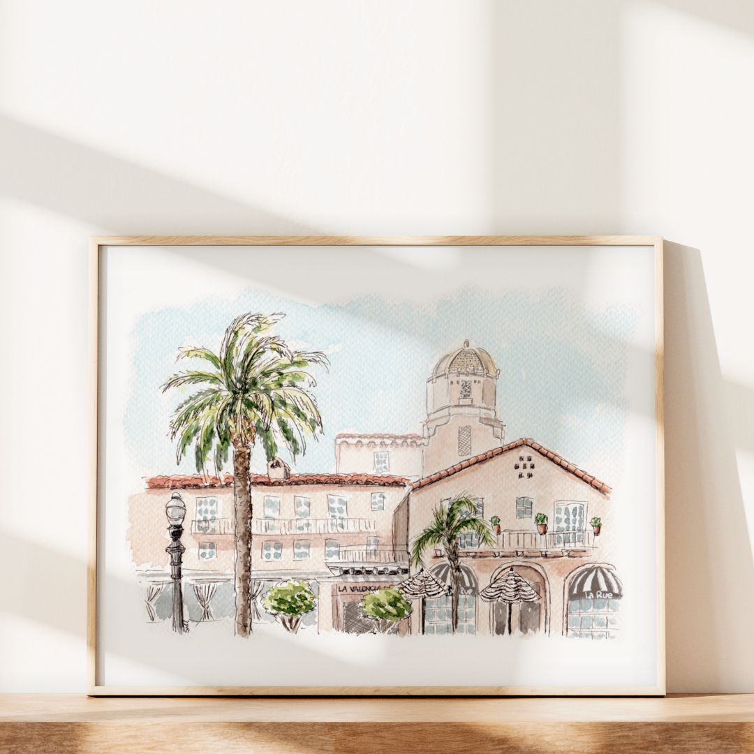 The Pink Lady of La Jolla by Swil Arts is framed watercolor wall art depicting a Mediterranean-style building with arched windows, striped awnings, a domed tower, and a palm tree, inspired by La Jolla’s iconic La Valencia Hotel.