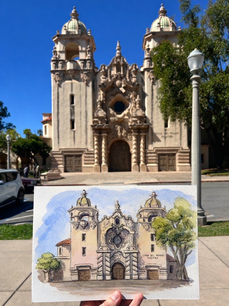 Real-life photograph of Teatro Casa del Prado, in Balboa Park, San Diego, with a hand holding a watercolor painting by Swil Arts of the same building.