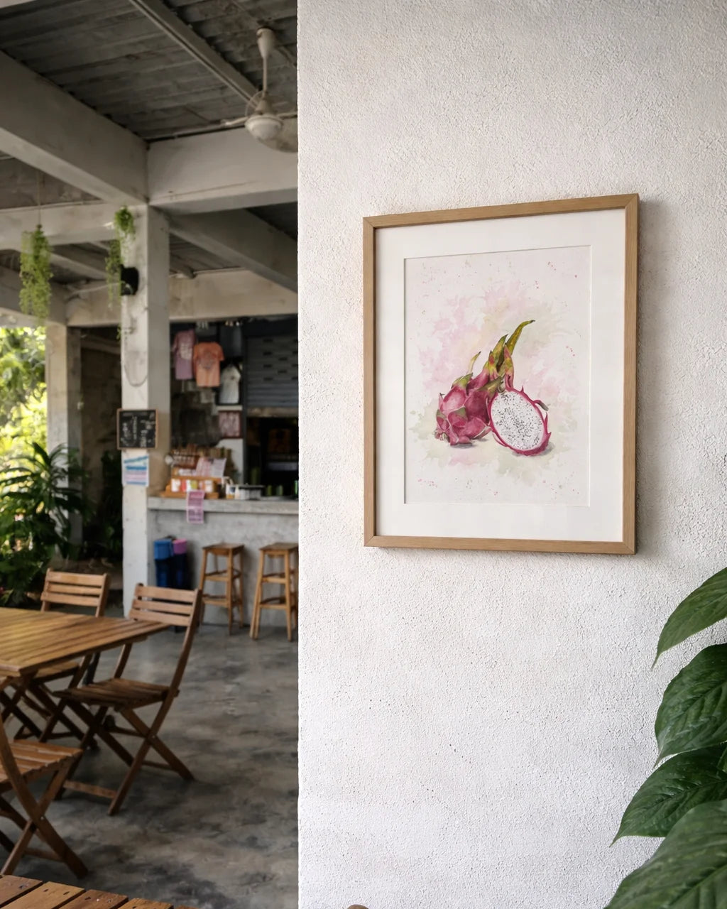 Framed artwork of a dragon fruit by Swil Arts, on a wall with wooden furniture and plants in the foreground.