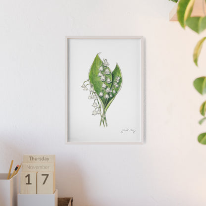 Framed botanical watercolor illustration of a lily of the valley flower with green leaves, signed by the artist 'Swil Arts,' displayed on a minimalist white wall. The artwork is styled in a light wood frame, next to a desktop setup featuring a wooden calendar showing 'Thursday, November 17,' and a pencil holder. A small plant adds a natural touch to the corner of the image.