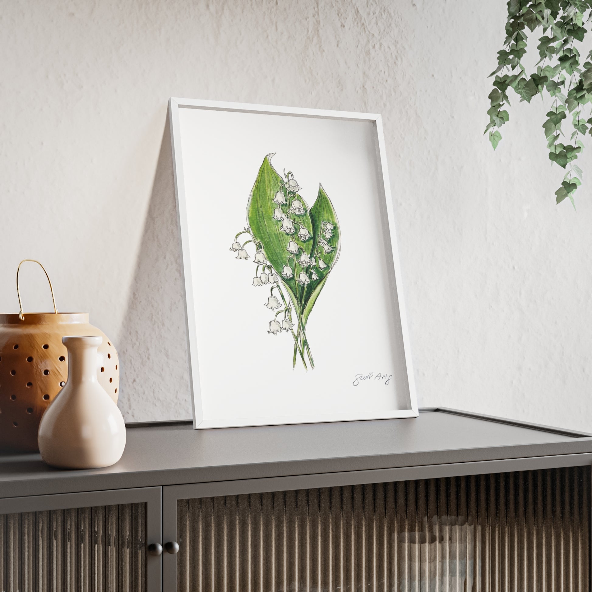 Framed botanical watercolor illustration of white lily of the valley flowers with green leaves, signed by the artist 'Swil Arts,' leaning against a white wall on top of a modern gray cabinet with ribbed glass doors. The setup includes a ceramic vase and a perforated lantern with a gold handle on the left, while a hanging ivy plant adds a touch of greenery to the scene. 