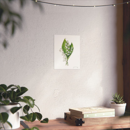 Lily of the Valley watercolor print by Swil Arts displayed in a cozy, minimalist interior. The delicate white flowers with green foliage are mounted on a textured wall, softly illuminated by natural light. A wooden surface below holds potted plants and books, creating a warm, nature-inspired aesthetic. Perfect for botanical art lovers and those seeking serene, eco-conscious home decor.