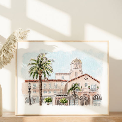 Pink Lady of La Jolla by Swil Arts—a watercolor wall art of a Mediterranean-style building with palm trees, inspired by the iconic La Valencia Hotel. Shown in a light wood frame with pampas grass and sunny window shadows beside it.