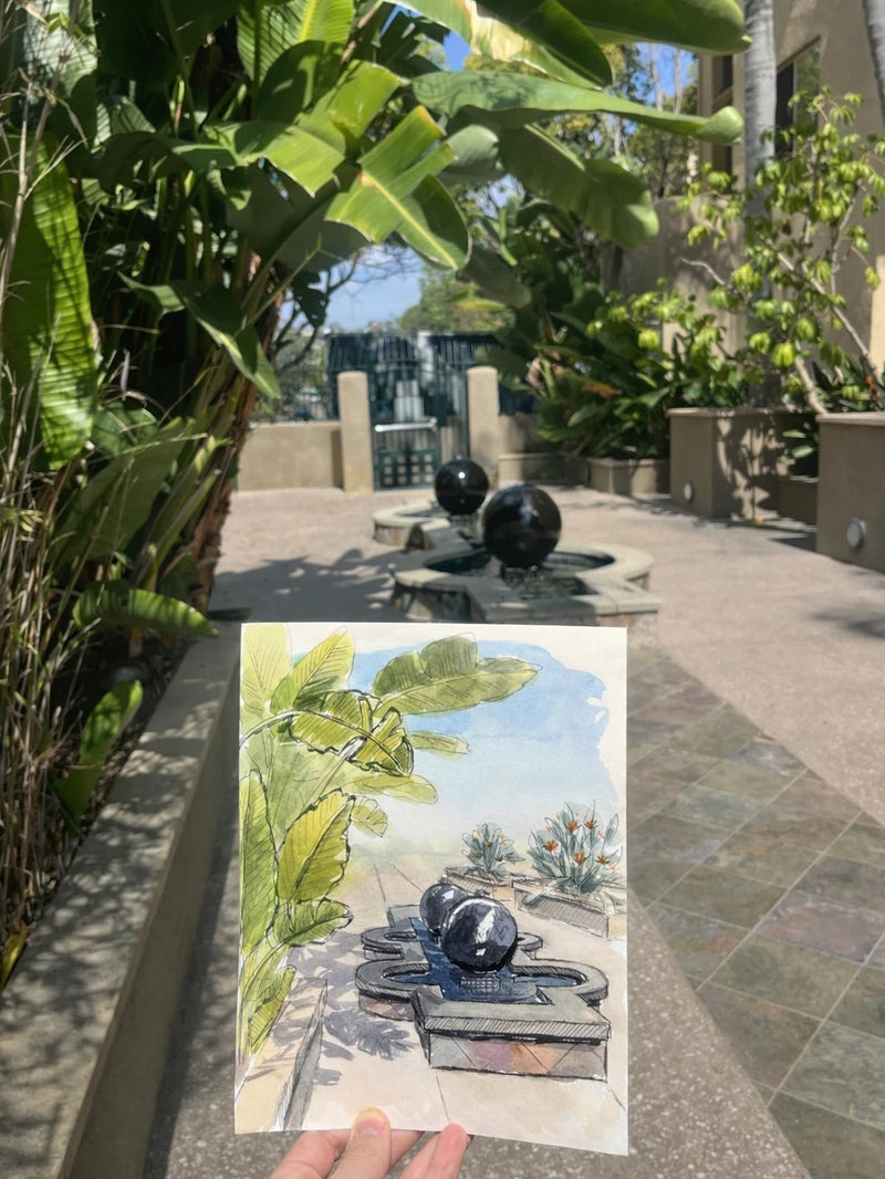 A hand holding a watercolor and ink painting of a garden scene in San Diego, with plants and a fountain.