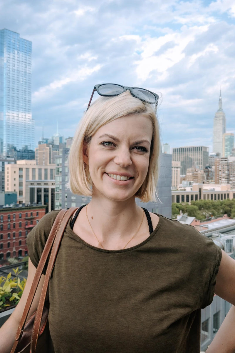 Sara, the artist behind Swil Arts, with platinum blond hair with sunglasses on head smiling in front of the New York City skyline