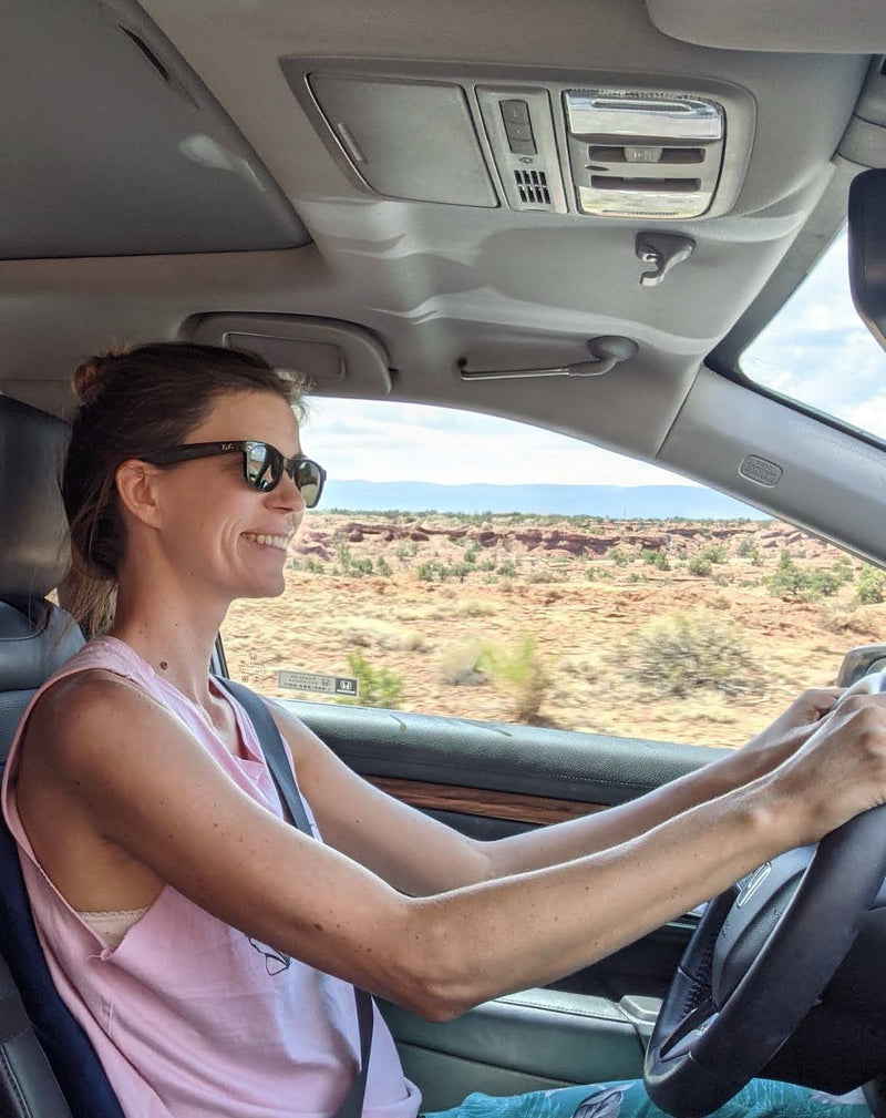 Sara, the artist behind Swil Arts, \ in a car driving through a desert landscape. Sara is wearing dark glasses and has a big smile on her face. 