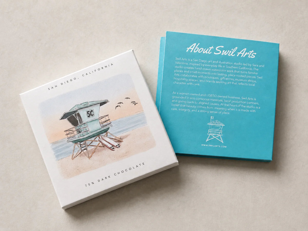 White chocolate box featuring a beach lifeguard painting by Sara from Swil Arts, next to a turquoise card cover insert with "About Swil Arts" text and logo on a beige surface.
