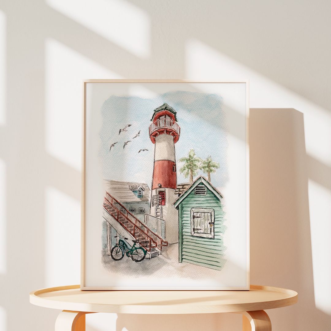The Lighthouse Tale by Swil Arts is a framed coastal watercolor showing a red and white lighthouse, green cottage, and bicycle among birds and palm trees, with sunlight casting soft shadows on the wall.