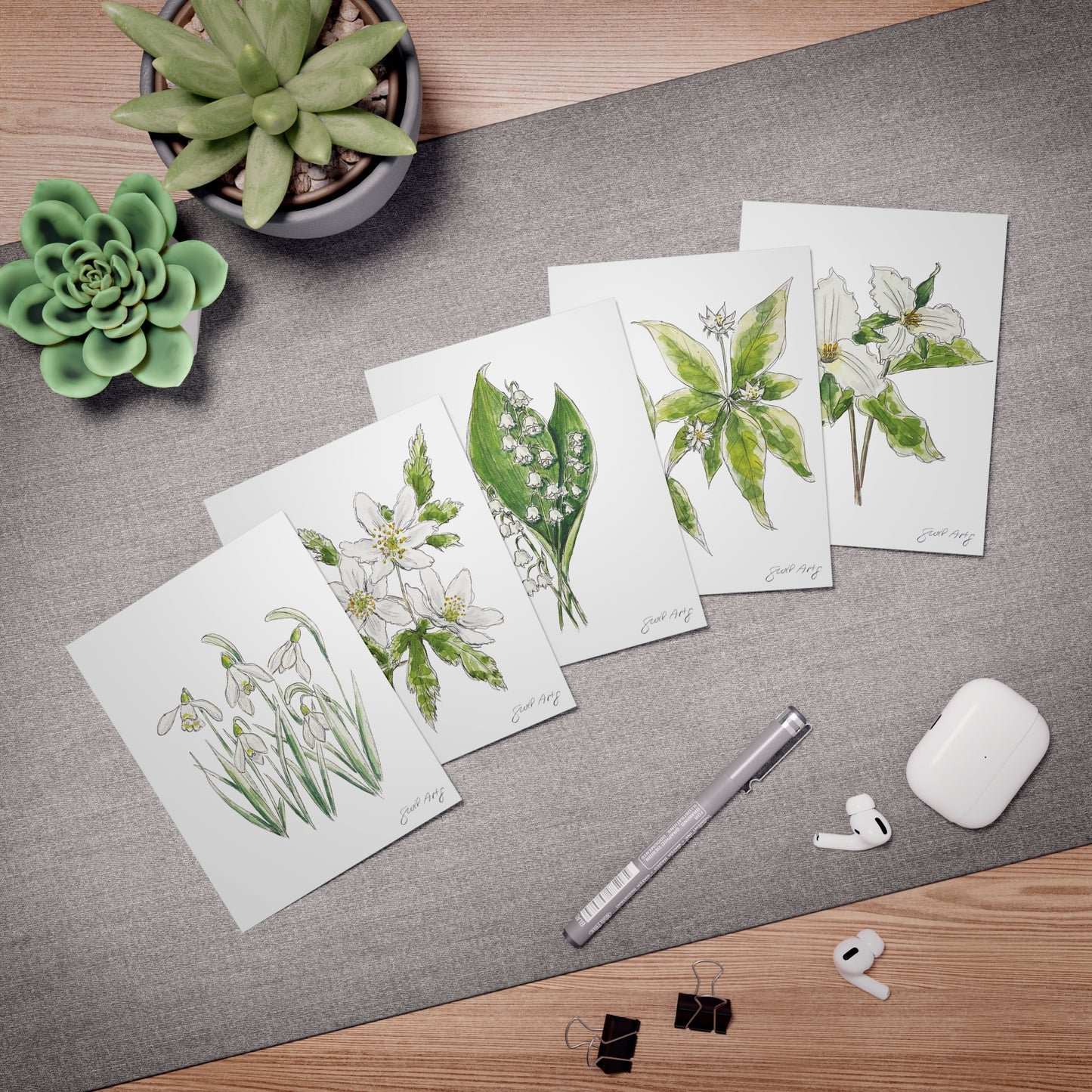 Set of botanical watercolor greeting cards by Swil Arts, featuring hand-illustrated floral designs. Each card showcases delicate ink and watercolor depictions of flowers, including snowdrops, anemones, lily of the valley, and trillium. Arranged on a textured gray surface with a fine liner pen, succulents, AirPods, and binder clips, these eco-friendly, artist-designed note cards are perfect for nature lovers, stationery enthusiasts, and heartfelt handwritten messages.
