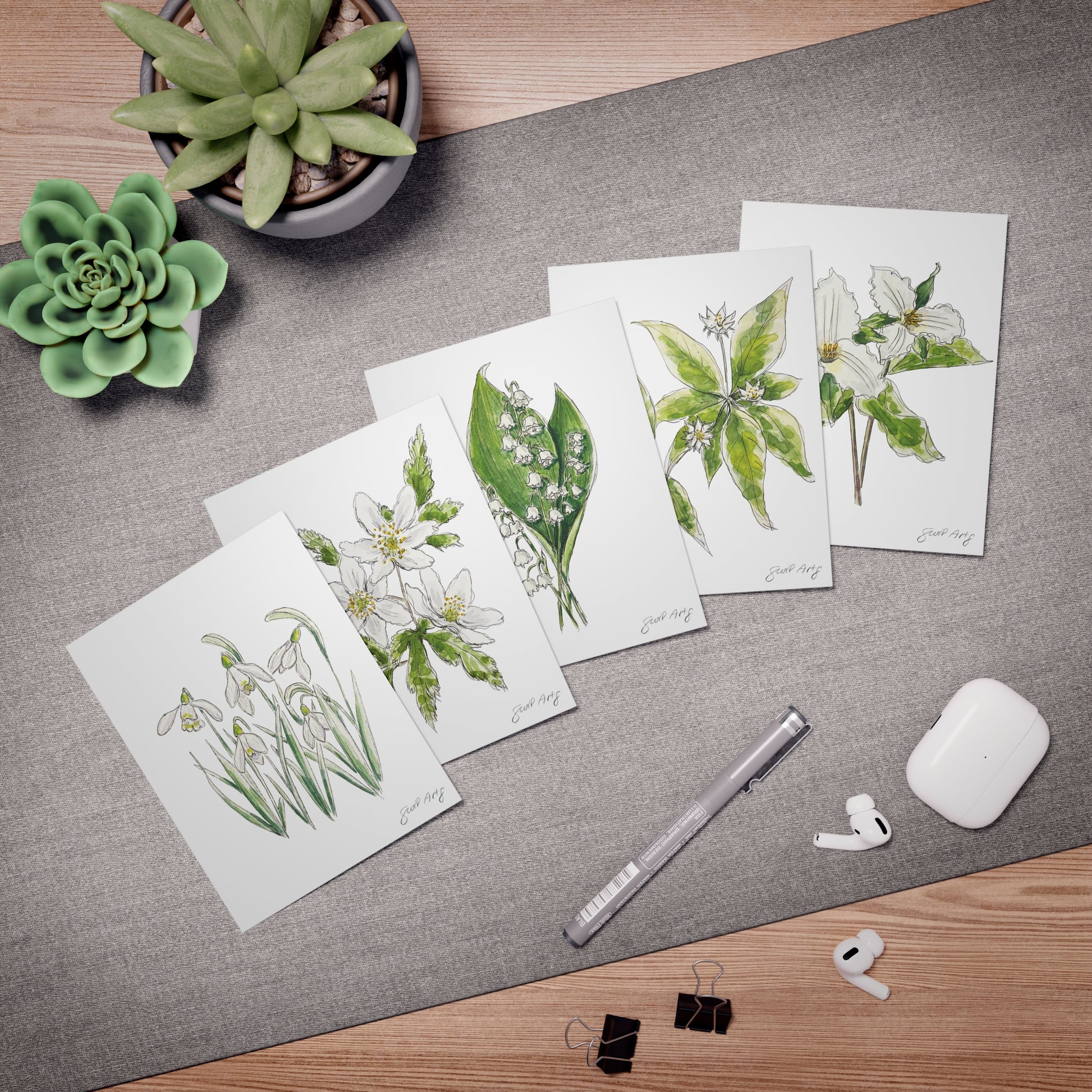 Set of botanical watercolor greeting cards by Swil Arts, featuring hand-illustrated floral designs. Each card showcases delicate ink and watercolor depictions of flowers, including snowdrops, anemones, lily of the valley, and trillium. Arranged on a textured gray surface with a fine liner pen, succulents, AirPods, and binder clips, these eco-friendly, artist-designed note cards are perfect for nature lovers, stationery enthusiasts, and heartfelt handwritten messages.