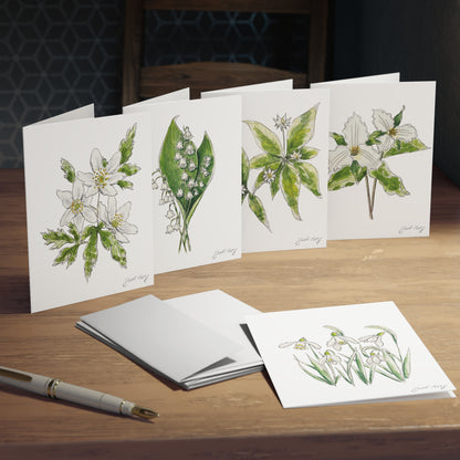 Set of botanical watercolor greeting cards by Swil Arts featuring delicate floral illustrations. Each card showcases hand-drawn flowers with intricate ink details and soft watercolor shading, including lily of the valley, trillium, and other nature-inspired designs. Displayed on a wooden table with envelopes and a fountain pen, these eco-friendly, artist-designed note cards are perfect for nature lovers, special occasions, and thoughtful handwritten messages.