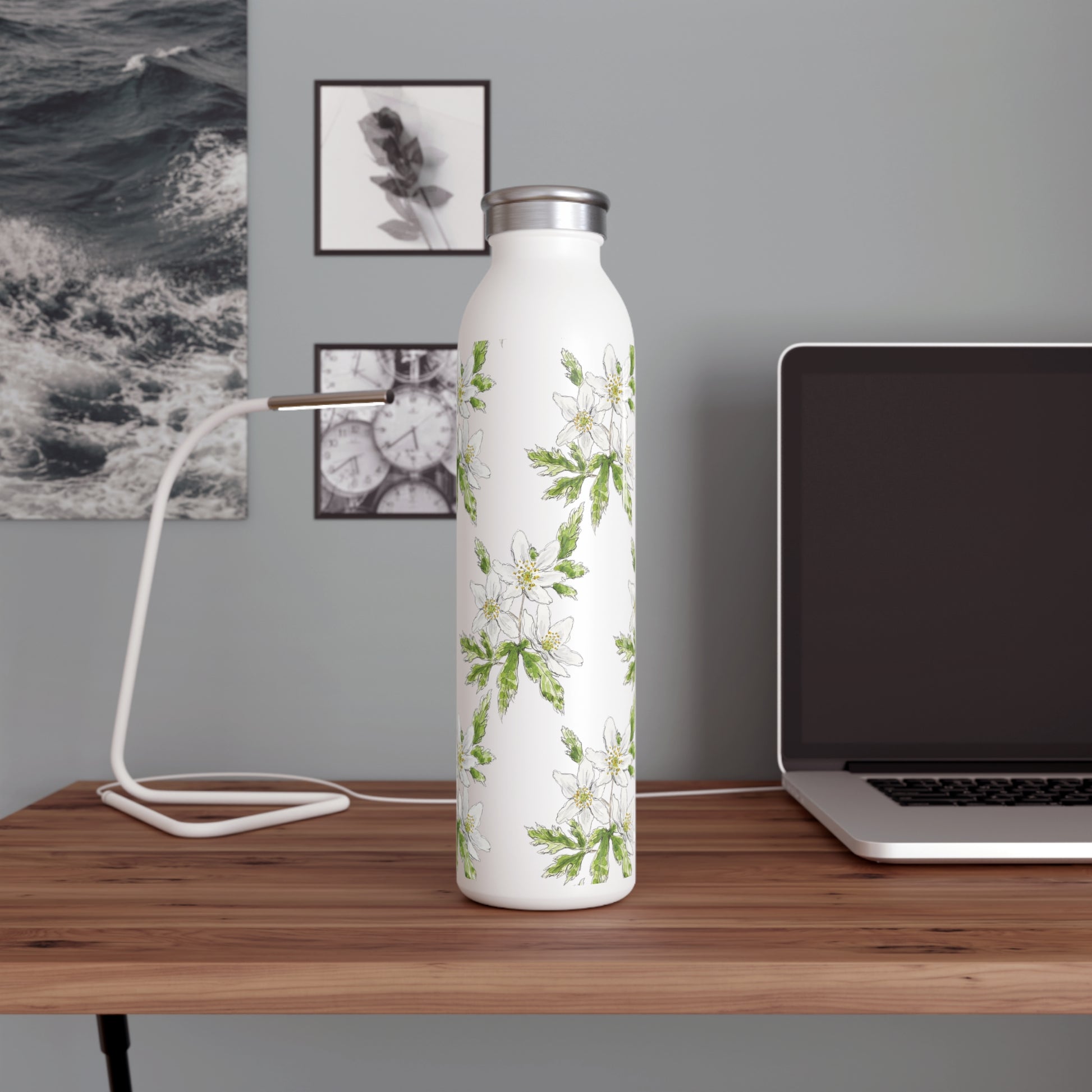 White stainless steel water bottle with a wood anemone flower watercolor design, placed on a wooden desk with a laptop, modern lamp, and black-and-white wall art in the background