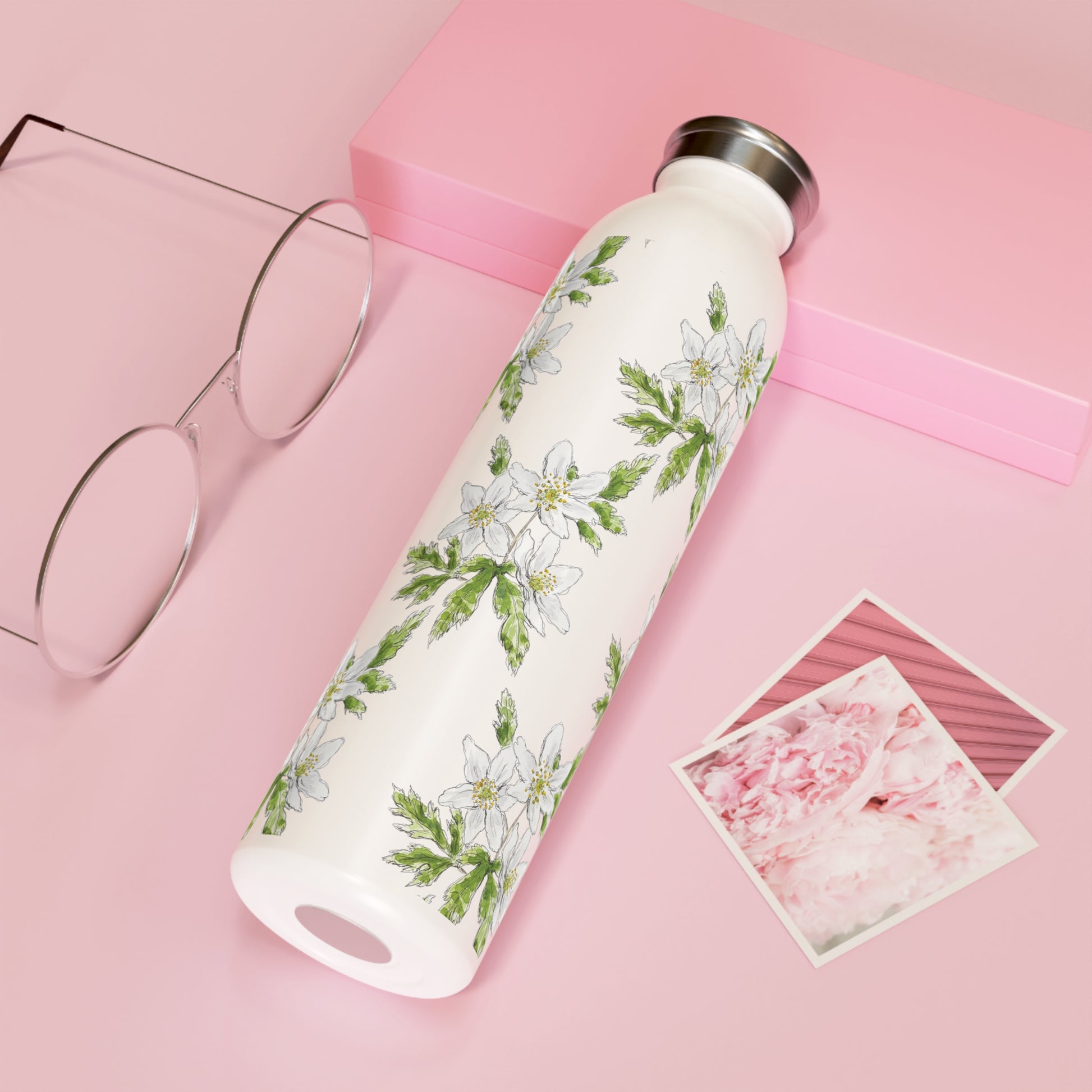 White stainless steel water bottle with a repeating wood anemone flower watercolor design, placed on a pink background with glasses and floral photographs.