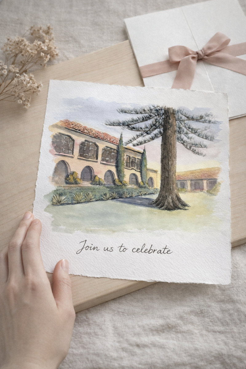 Handheld card with a watercolor and ink illustration of Liberty Station wedding venue and invitation text, surrounded by gift boxes and flowers on a textured surface.