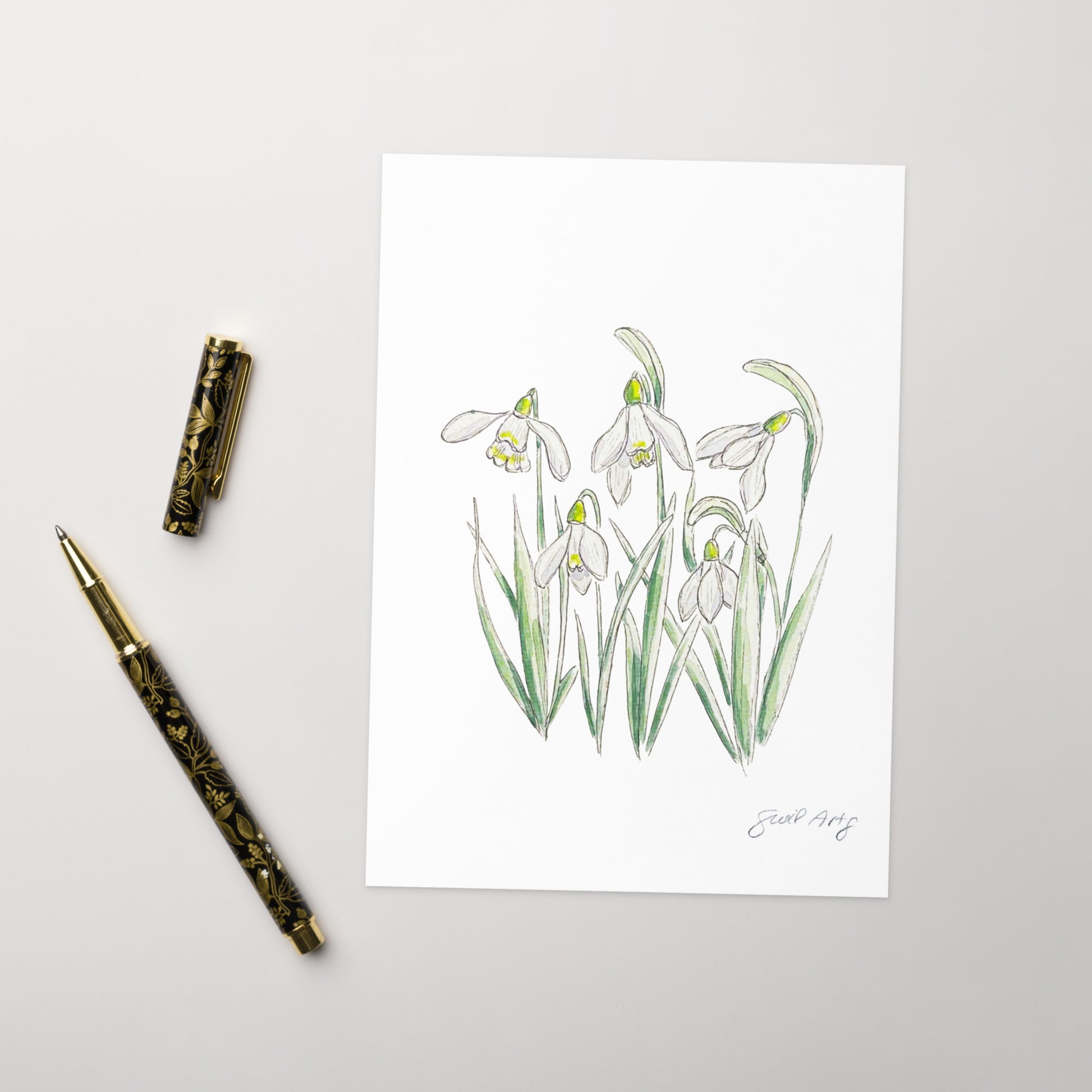Snowdrop flower greeting card with a delicate watercolor and ink illustration. Botanical note card featuring a woodland-inspired floral design, perfect for nature lovers. The greeting card is nicely set with an elegant gold en next to it, ready to journal.