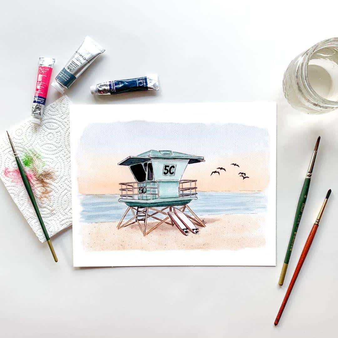 Watercolor painting of a Southern California lifeguard tower at sunset, accompanied by paint tubes, brushes, and a jar of water. Handcrafted coastal artwork in progress by Swil Arts