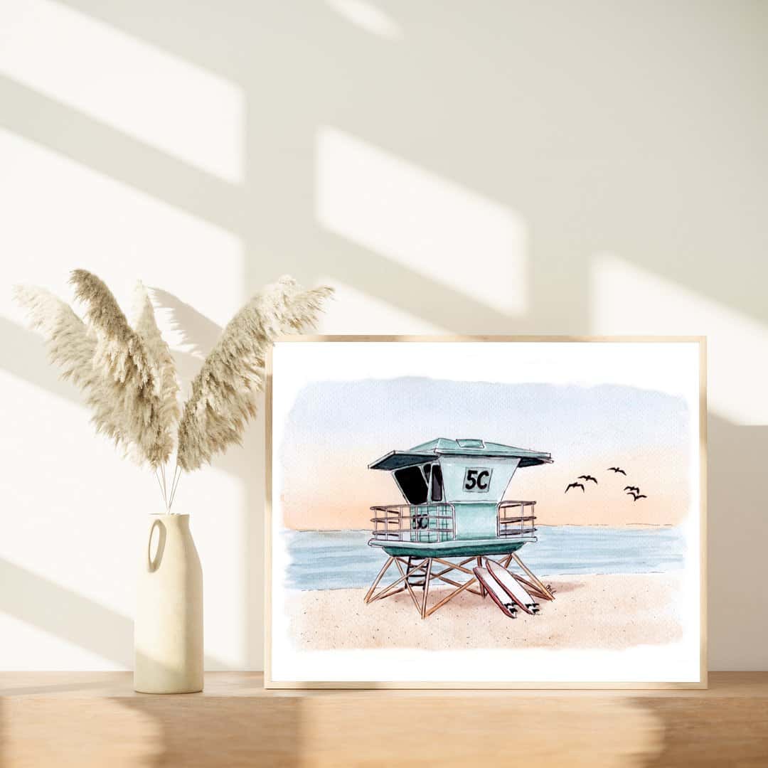 Framed watercolor and ink print of a Southern California lifeguard tower at sunset, featuring surfboards and birds in flight, displayed next to a vase with pampas grass. Handmade coastal decor by Swil Arts