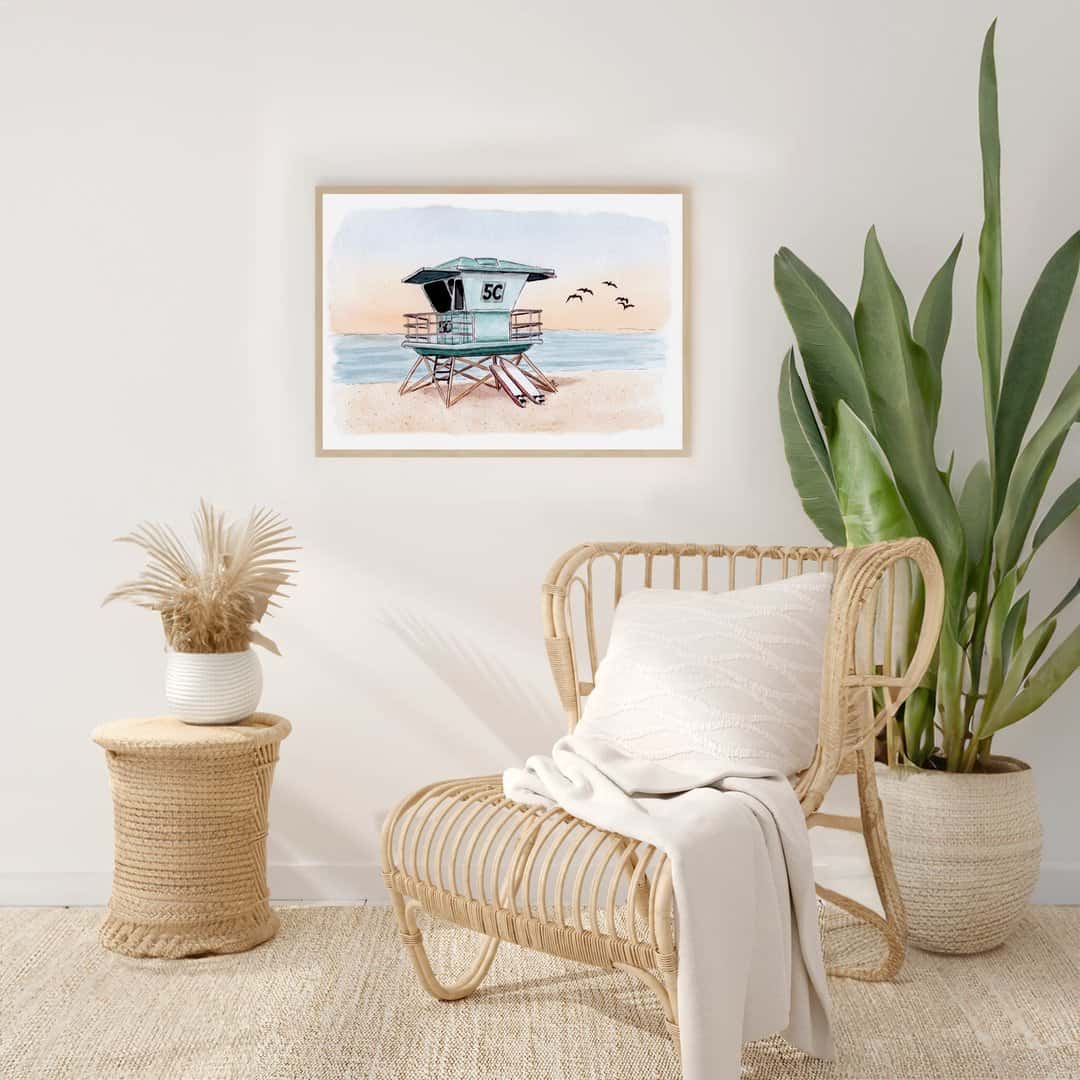 Framed watercolor and ink print of a Southern California lifeguard tower at sunset with surfboards leaning against the tower, displayed in a modern coastal living room setting. Handmade artwork by Swil Arts, perfect for beach lovers and coastal decor enthusiasts.