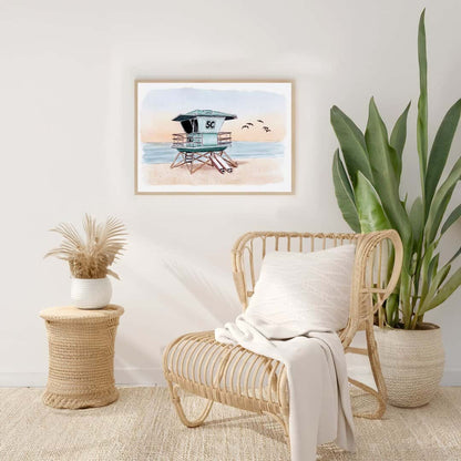 Framed watercolor and ink print of a Southern California lifeguard tower at sunset with surfboards leaning against the tower, displayed in a modern coastal living room setting. Handmade artwork by Swil Arts, perfect for beach lovers and coastal decor enthusiasts.