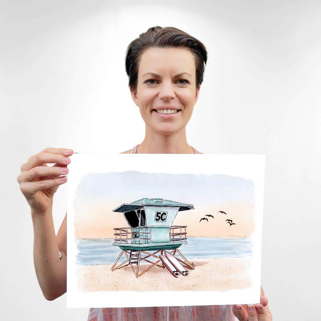 Sara, founder and artist of Swil Arts, smiling proudly while holding her handmade watercolor and ink print of a Southern California lifeguard tower at sunset, with surfboards leaning against the tower.