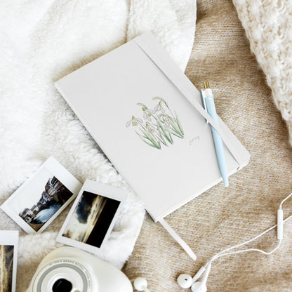 White hardcover notebook featuring a snowdrop flower illustration by Swil Arts, resting on a cozy textured blanket with a pen, instant film photos, a vintage camera, and white earbuds. Aesthetic stationery for journaling, writing, and creative inspiration.