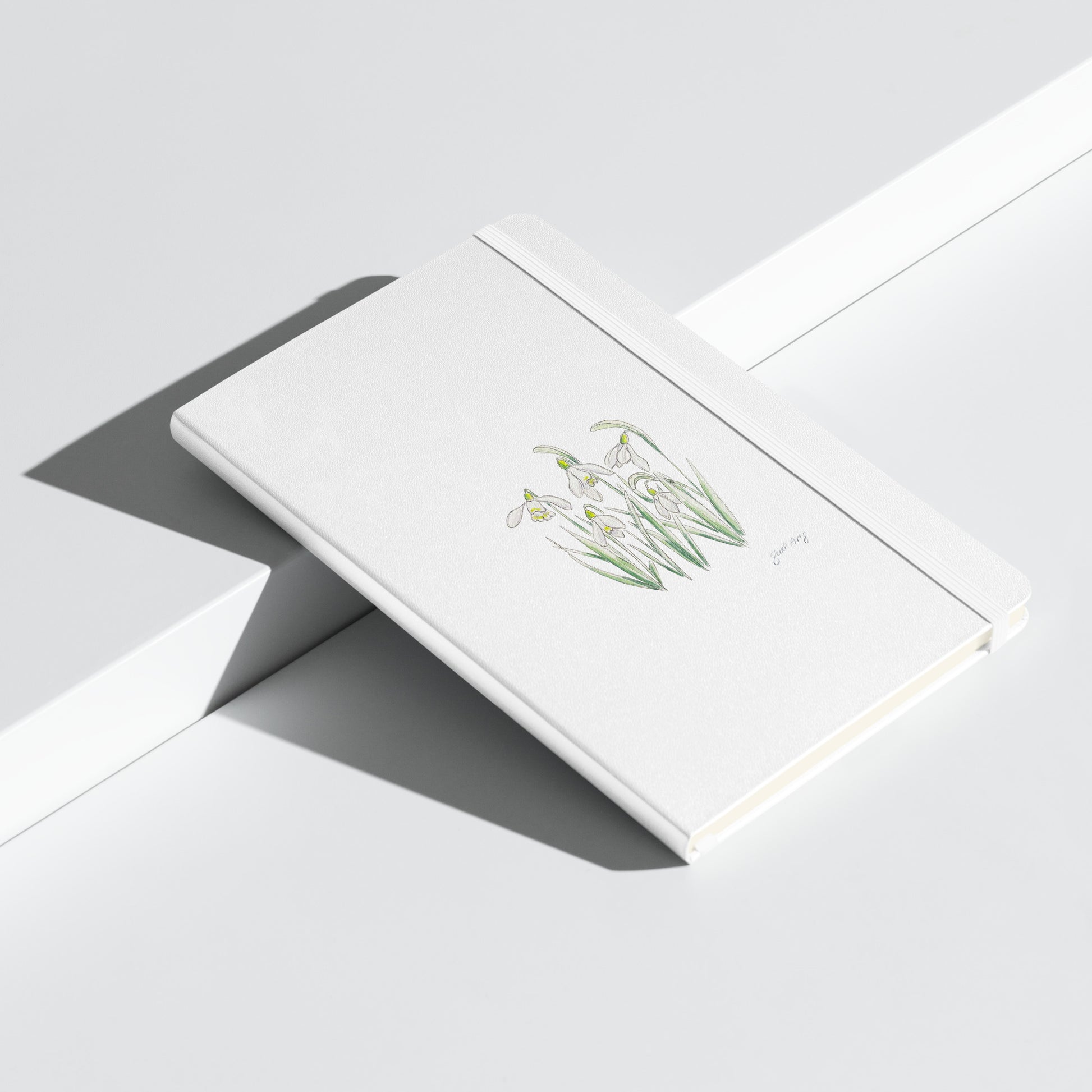 White hardcover notebook featuring a delicate snowdrop flower illustration by Swil Arts. This botanical watercolor and ink design showcases the beauty of nature, making it perfect for journaling, sketching, or note-taking. The notebook includes an elastic closure for convenience, ideal for artists, writers, and nature lovers seeking elegant stationery with a floral touch.