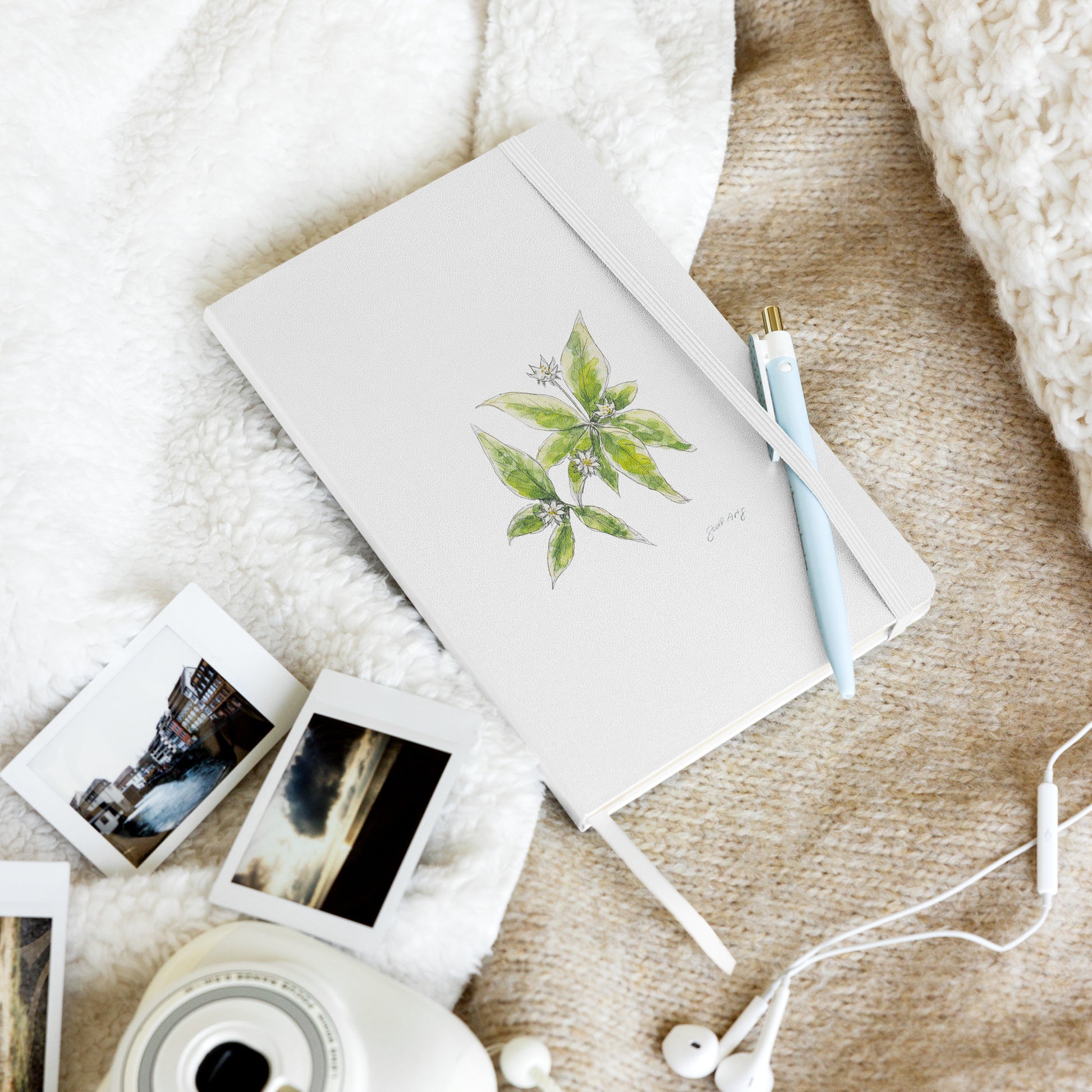 White hardcover notebook featuring a  starflower illustration by Swil Arts, resting on a cozy textured blanket with a pen, instant film photos, a vintage camera, and white earbuds. Aesthetic stationery for journaling, writing, and creative inspiration.