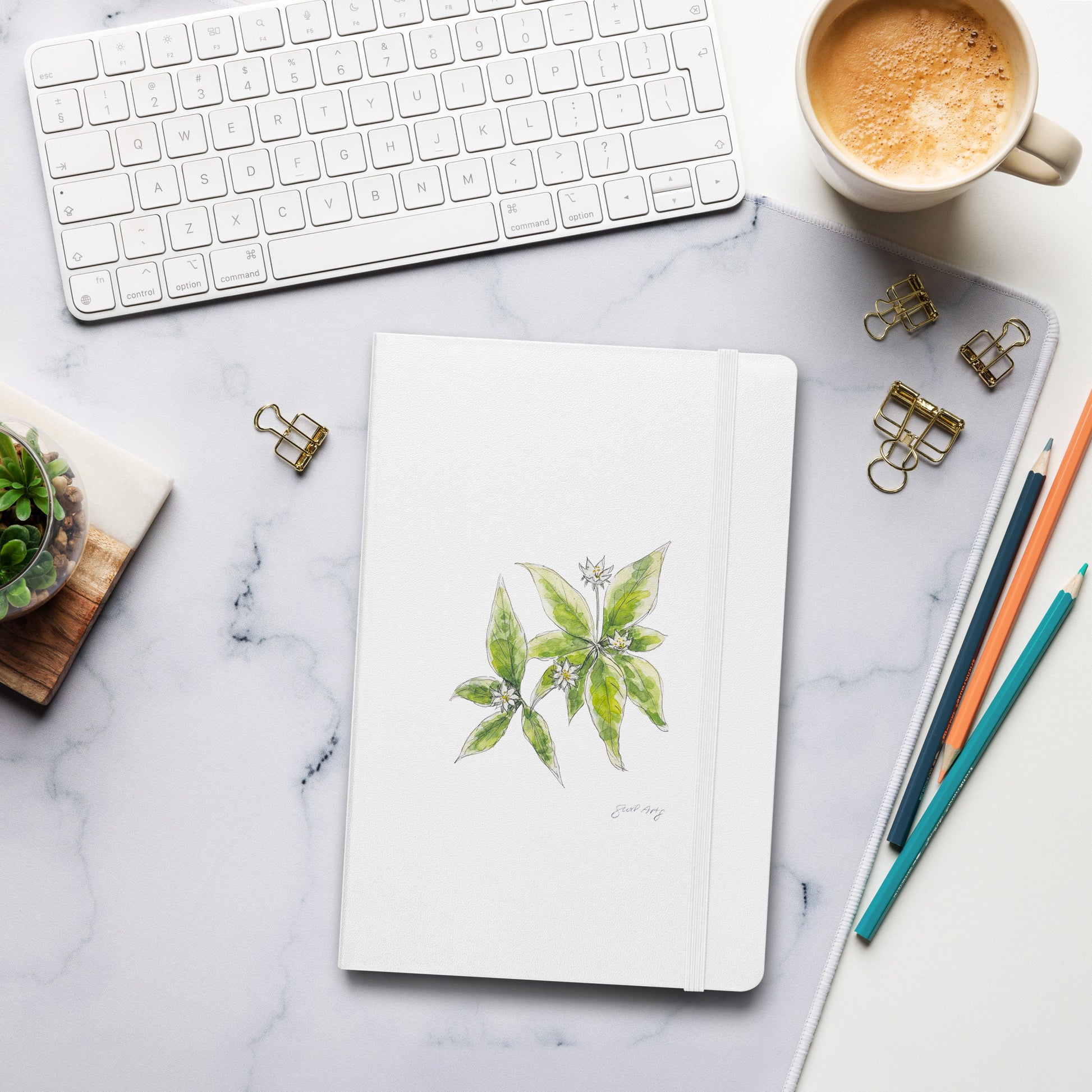 White hardcover notebook with a starflower illustration by Swil Arts, placed on a marble desk with a keyboard, coffee, pencils, and gold binder clips. Aesthetic stationery for journaling, writing, and sketching.