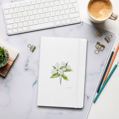 White hardcover notebook with a trillium flower illustration by Swil Arts, placed on a marble desk with a keyboard, coffee, pencils, and gold binder clips. Aesthetic stationery for journaling, writing, and sketching.