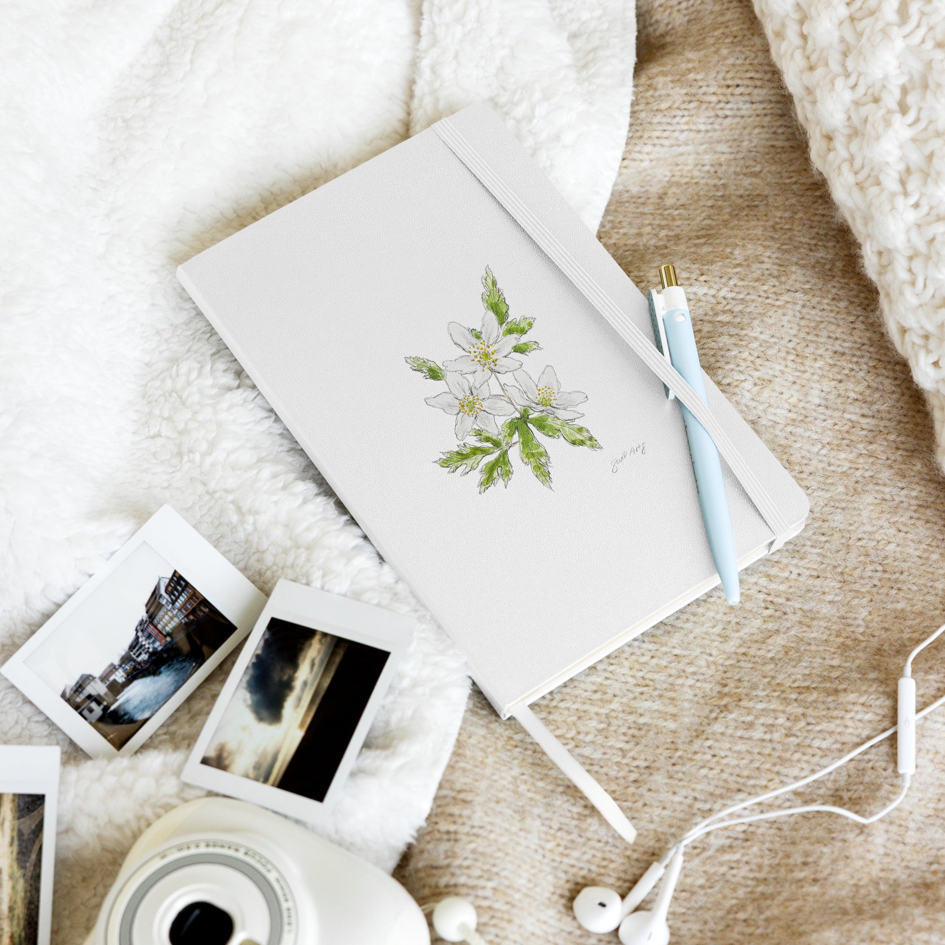 White hardcover notebook featuring a wood anemone flower illustration by Swil Arts, resting on a cozy textured blanket with a pen, instant film photos, a vintage camera, and white earbuds. Aesthetic stationery for journaling, writing, and creative inspiration.