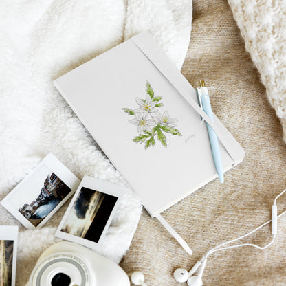 White hardcover notebook featuring a wood anemone flower illustration by Swil Arts, resting on a cozy textured blanket with a pen, instant film photos, a vintage camera, and white earbuds. Aesthetic stationery for journaling, writing, and creative inspiration.