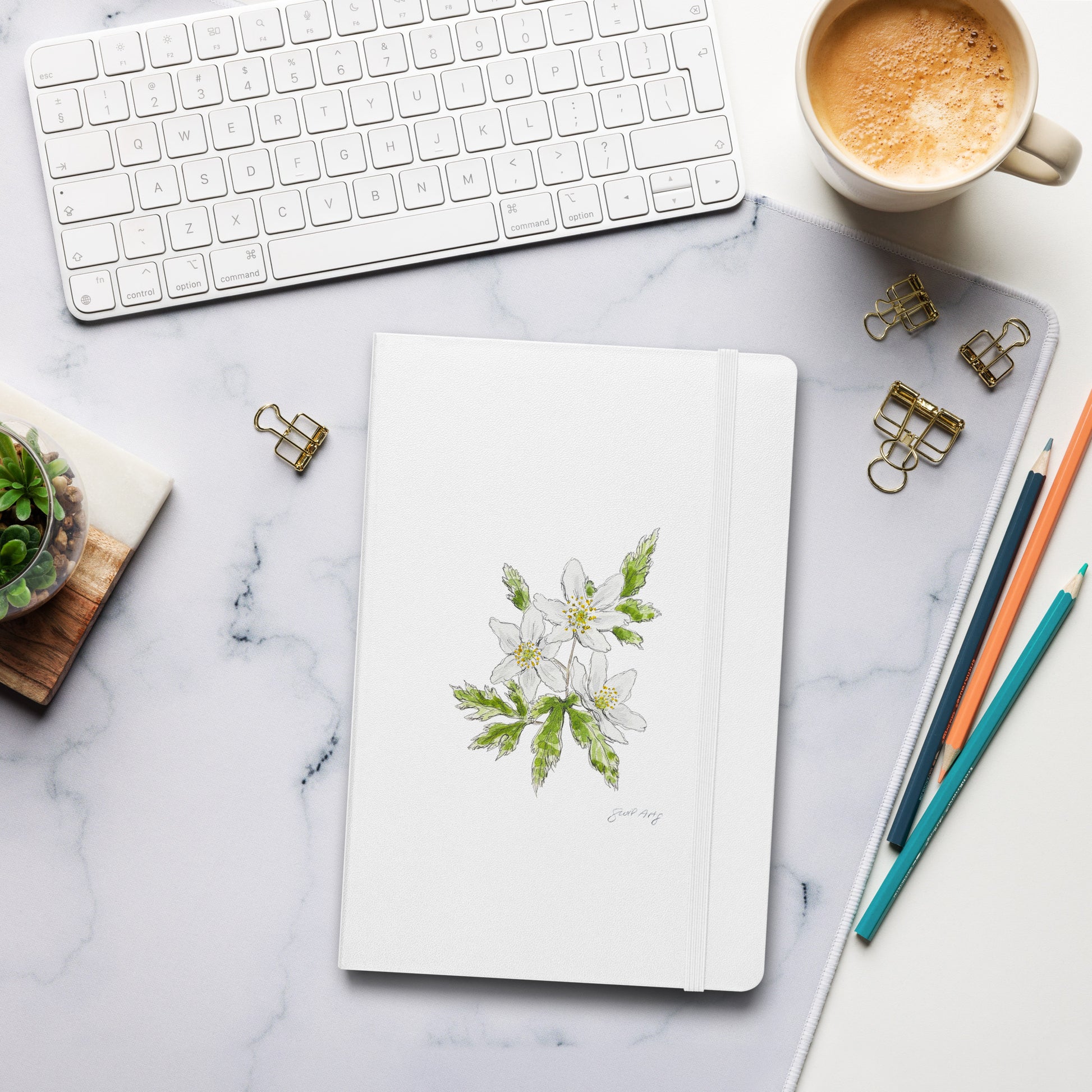 White hardcover notebook with a wood anemone flower illustration by Swil Arts, placed on a marble desk with a keyboard, coffee, pencils, and gold binder clips. Aesthetic stationery for journaling, writing, and sketching.