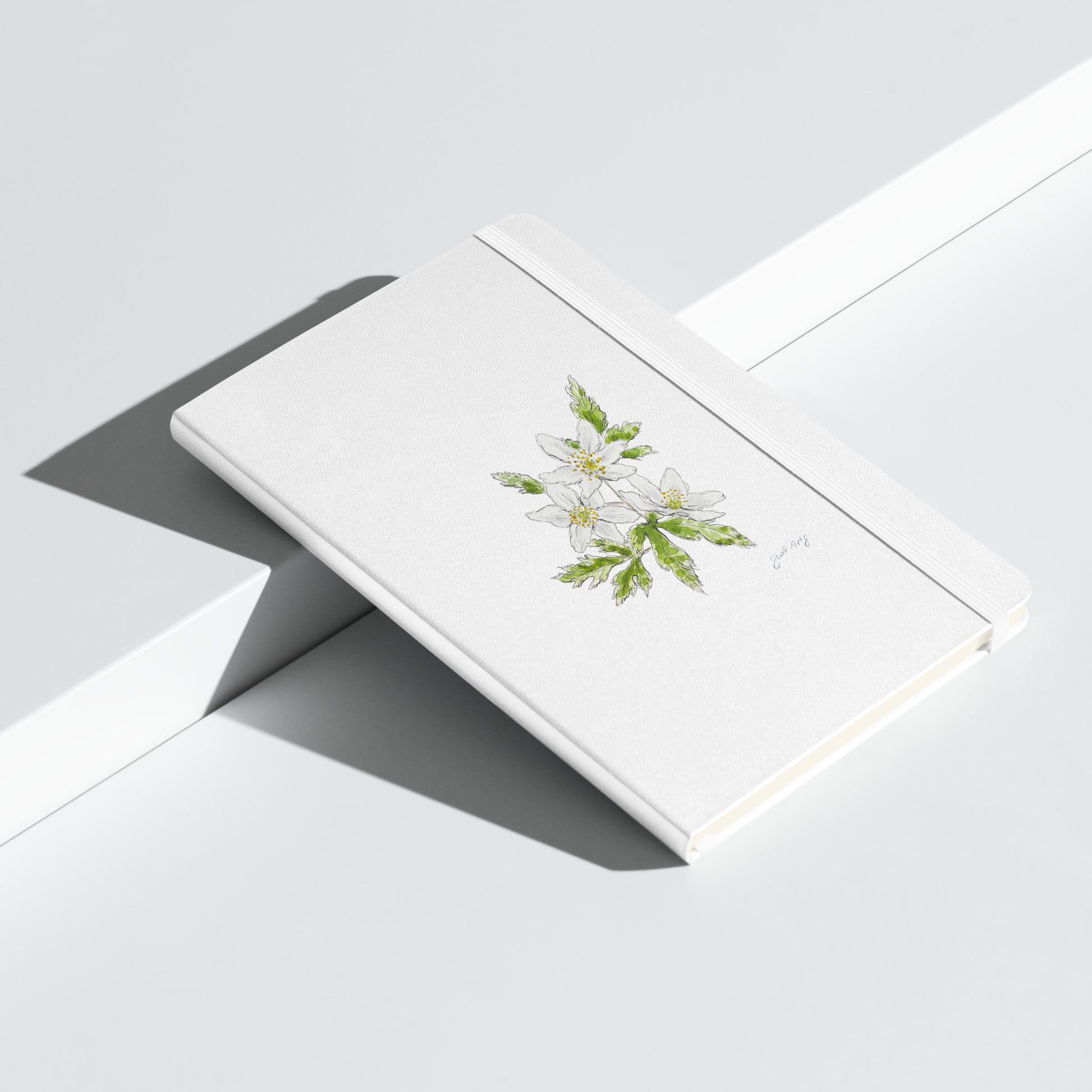 White hardcover notebook featuring a delicate wood anemone flower illustration by Swil Arts. This botanical watercolor and ink design showcases the beauty of nature, making it perfect for journaling, sketching, or note-taking. The notebook includes an elastic closure for convenience, ideal for artists, writers, and nature lovers seeking elegant stationery with a floral touch.