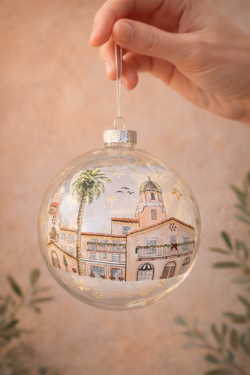 Decorative glass ornament with a painted scene of a La Valencia Hotel in La Jolla and palm tree, held by a hand against a beige background.