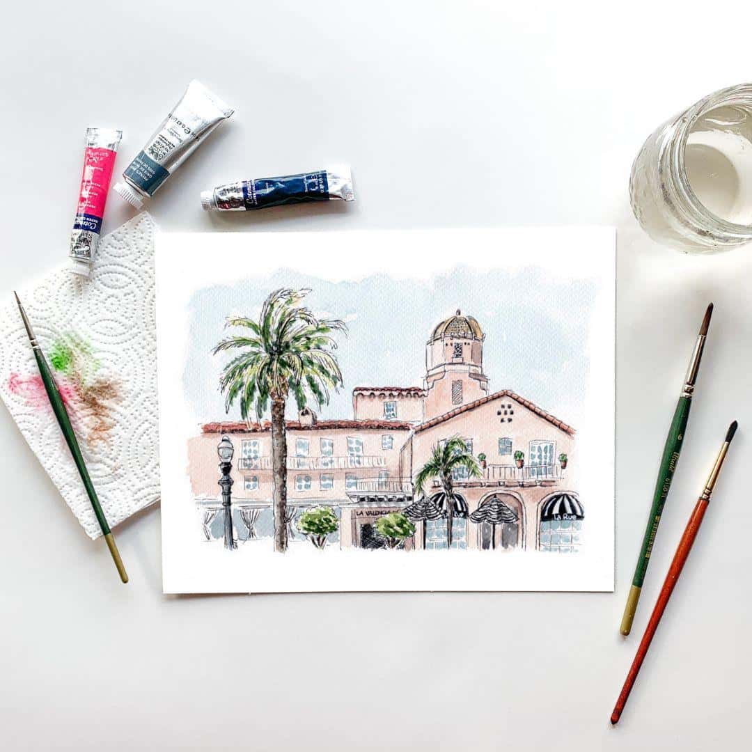 Watercolor painting of La Valencia Hotel in La Jolla, California, with palm trees and Mediterranean-inspired architecture. The pink-hued building features a dome tower and arched windows, painted in soft pastel tones. Surrounded by artist tools like paintbrushes, watercolor tubes, and a water jar on a white background