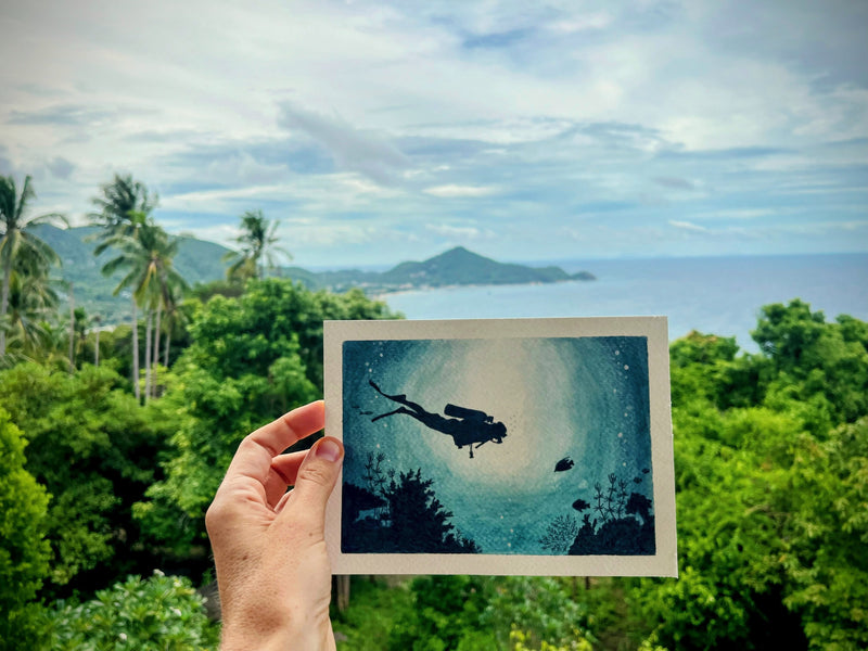 Hand holding a framed artwork of a diver underwater with a scenic ocean view in the background.
