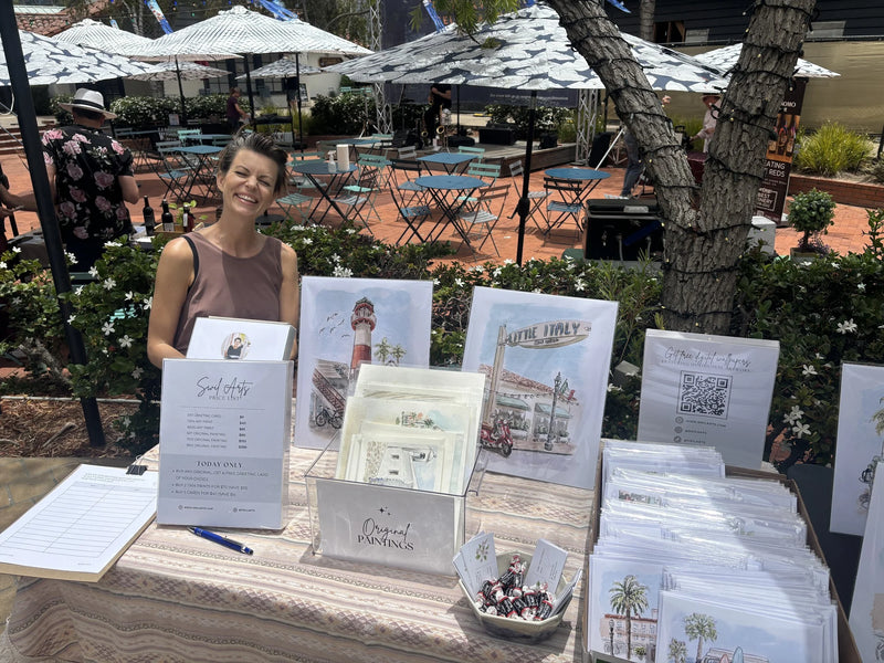 Sara from Swil Arts stands behind a table displaying printed wedding stationery and cards in an outdoor patio setting with umbrellas, tables, and trees in the background.