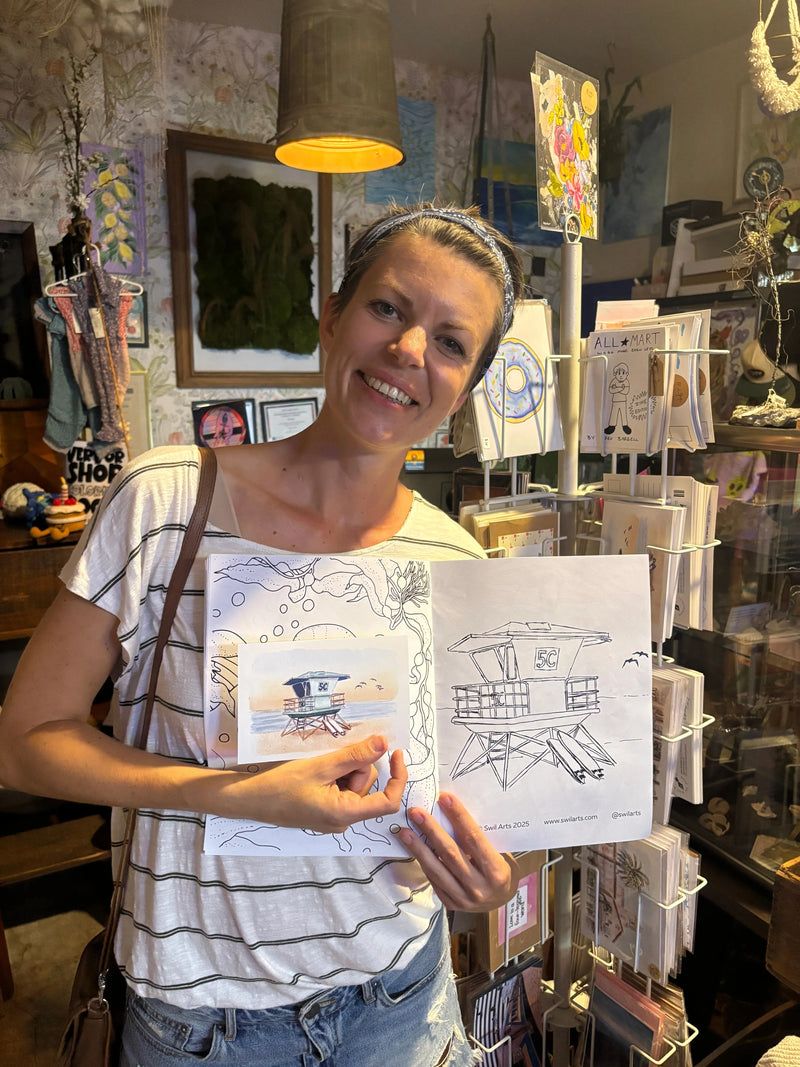 Sara from Swil Arts, smiling, in a white and black striped shirt holds up a sketchbook with architectural line drawings and a small color illustration, set against a busy shop background filled with art supplies and framed pieces.