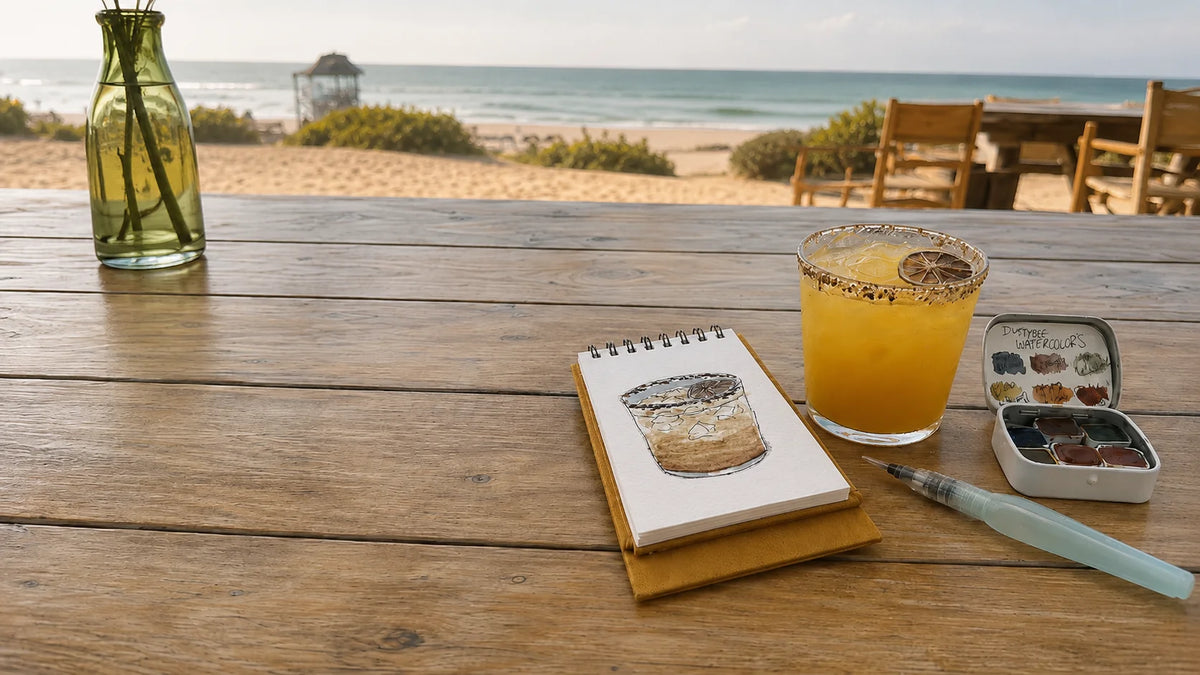 A styled beach-side tabletop scene with a spiral notebook showing a sketch of a cocktail, a glass of orange drink with a lime wedge, a small paint palette, and a paintbrush on a wooden table, with a beach and ocean in the background. Artwork by Sara from Swil Arts