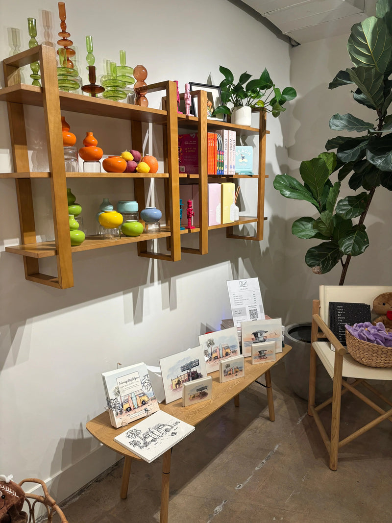 A styled interior display featuring light wood shelving units with colorful decorative objects, a white table holding printed cards or brochures, and a large potted green plant in a modern room with a concrete floor. Artwork by Sara from Swil Arts