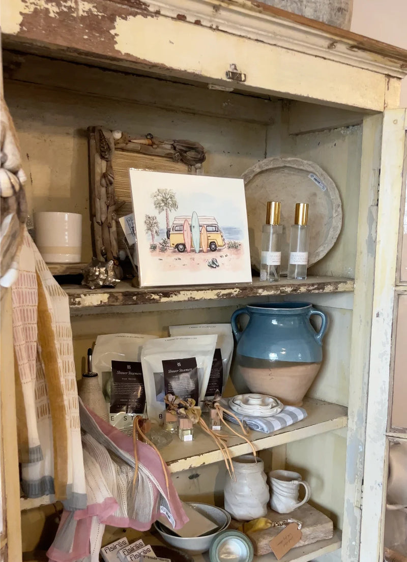 A rustic, distressed cream-colored wooden cabinet or shelf display filled with various home decor items, including a small watercolor art print of a camper van, a blue ceramic vase, glass bottles, and other decorative accessories. Artwork by Sara from Swil Arts