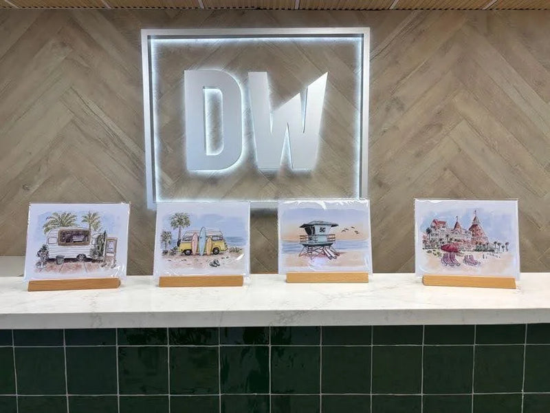 Four small framed art prints with beach and travel-themed illustrations are displayed on a white countertop in front of a large glowing 'DW' sign mounted on a wood-paneled wall. Artwork by Sara from Swil Arts