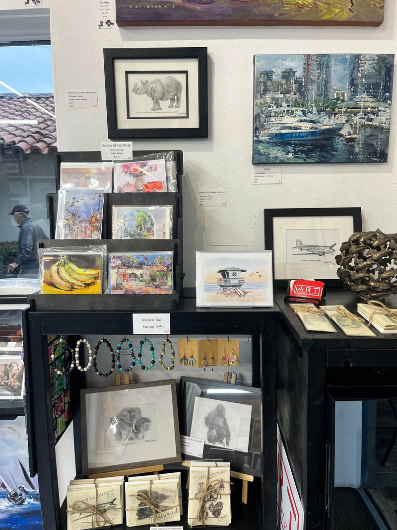 A retail display shelf showcasing a variety of art prints, framed sketches, greeting cards, and jewelry, set against a white wall with other framed artwork in the background. You can see Guardian of the Shores painted by Sara from Swil Arts in the middle