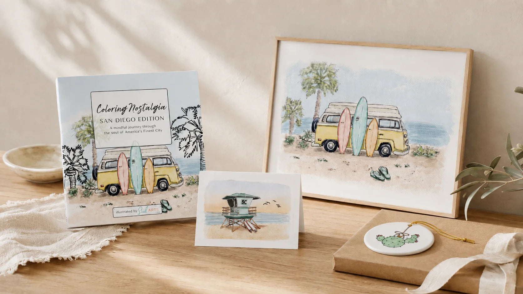 A styled flat-lay of a coloring book titled 'Coloring Nostalgia: San Diego Edition' next to a framed watercolor art print of a yellow camper van on a beach, with a small card, a wrapped ornament, and a lace cloth on a wooden surface. Artwork by Sara from Swil Arts