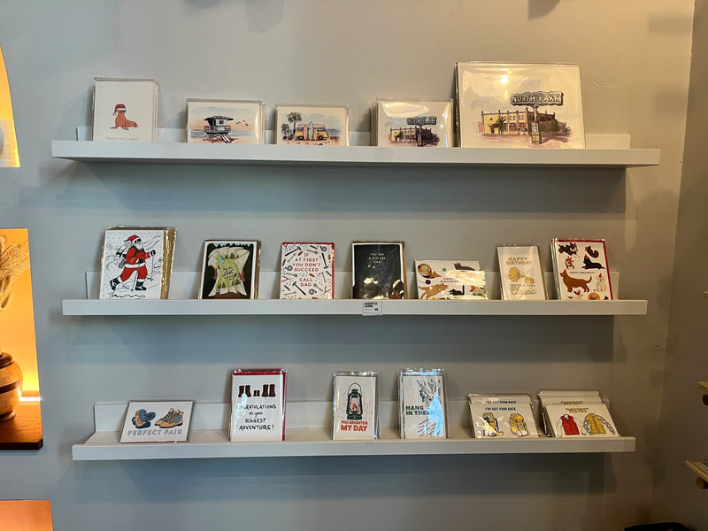 Three white floating shelves mounted on a light gray wall, displaying a curated collection of small framed art prints, cards, and books with vintage-style illustrations. Artwork by Sara from Swil Arts