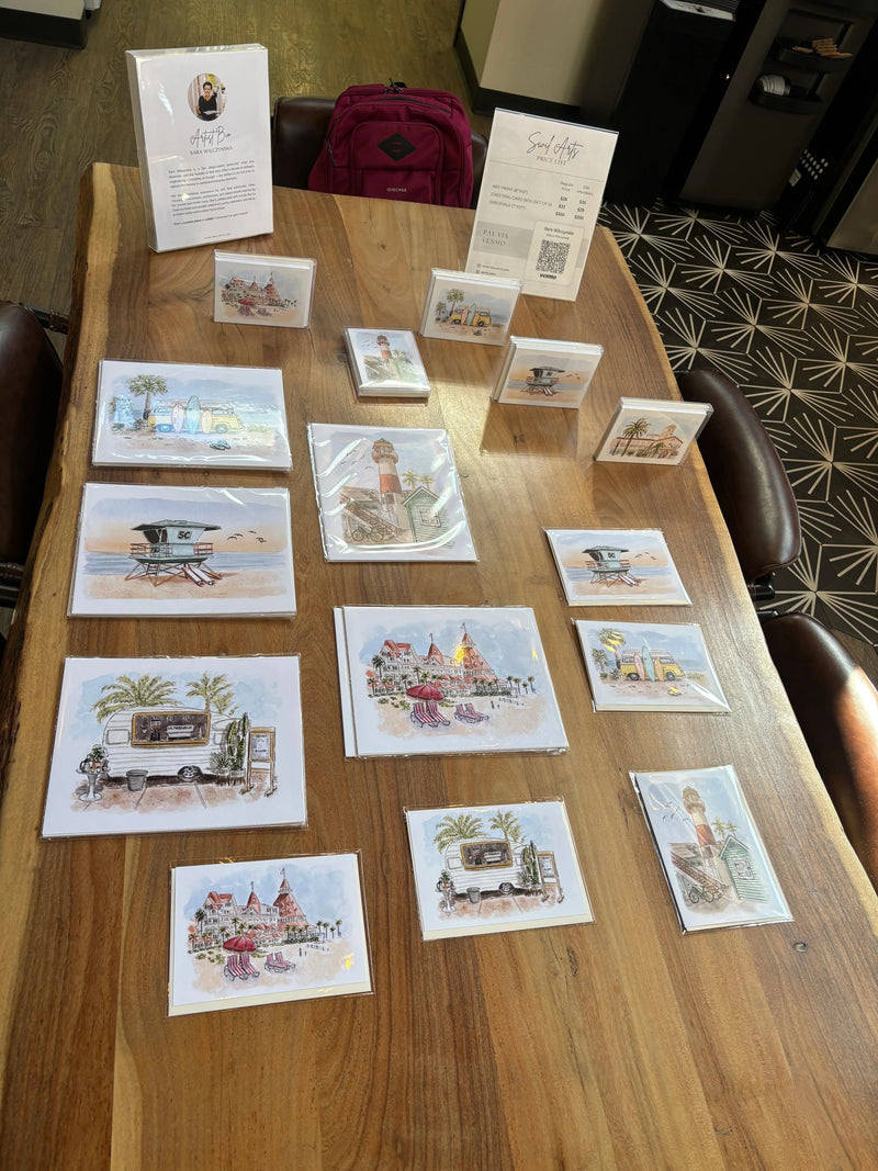 A collection of small illustrated art prints and postcards displayed on a wooden table, with some cards standing upright and others lying flat. The prints feature colorful scenic and architectural illustrations. Artwork by Sara from Swil Arts